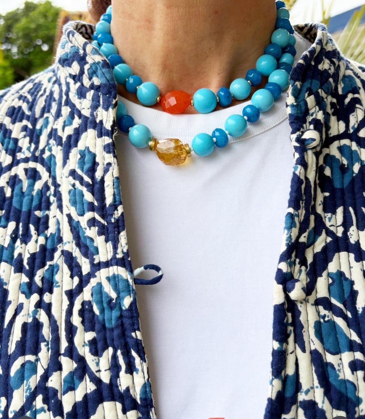 Collar corto artesanal de piedras semipreciosas azules, naranja y amarilla sobre camisa blanca y chaqueta azul con estampado .